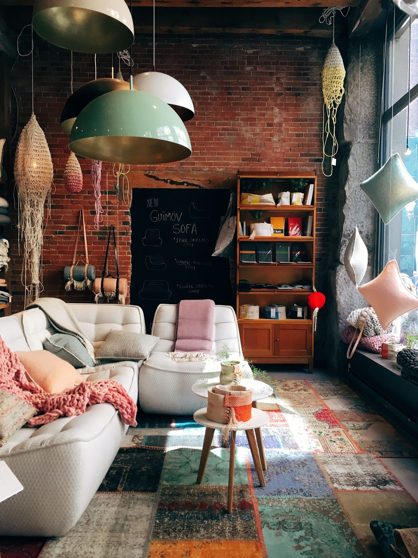 An inviting room with a brick back wall and a large window to the right. A rug with multi-coloured rectangle pattern is on the floor. A large, white sofa adorned with pink and grey cushions faces the window. Large metal lamps hang over the sofa. A wooden bookcase with boxes and notebooks is against the back wall, along with a blackboard.