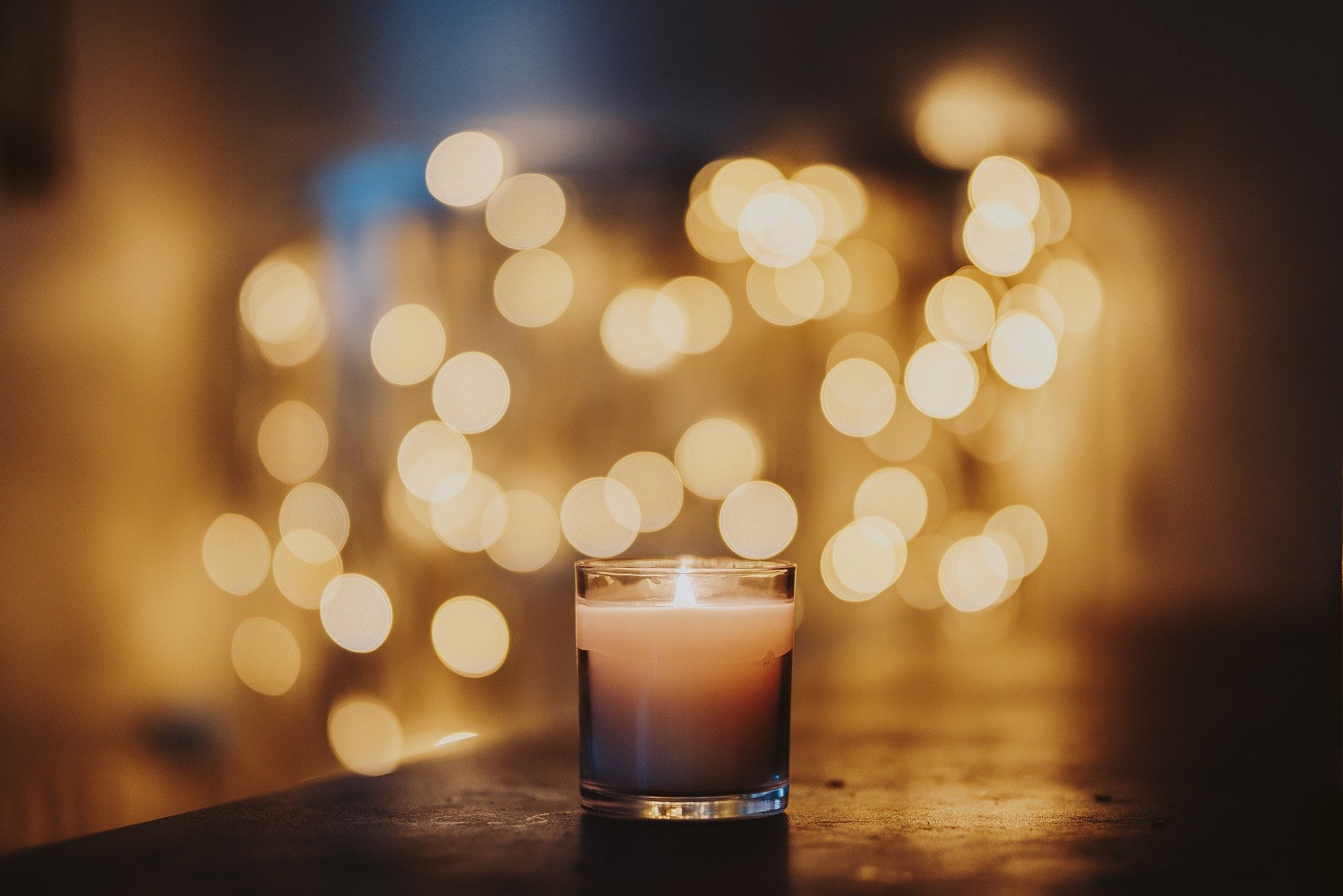 A small, lit peach-coloured candle in a glass on a table in a dark room. In the background, fairy lights glow a warm yellow.
