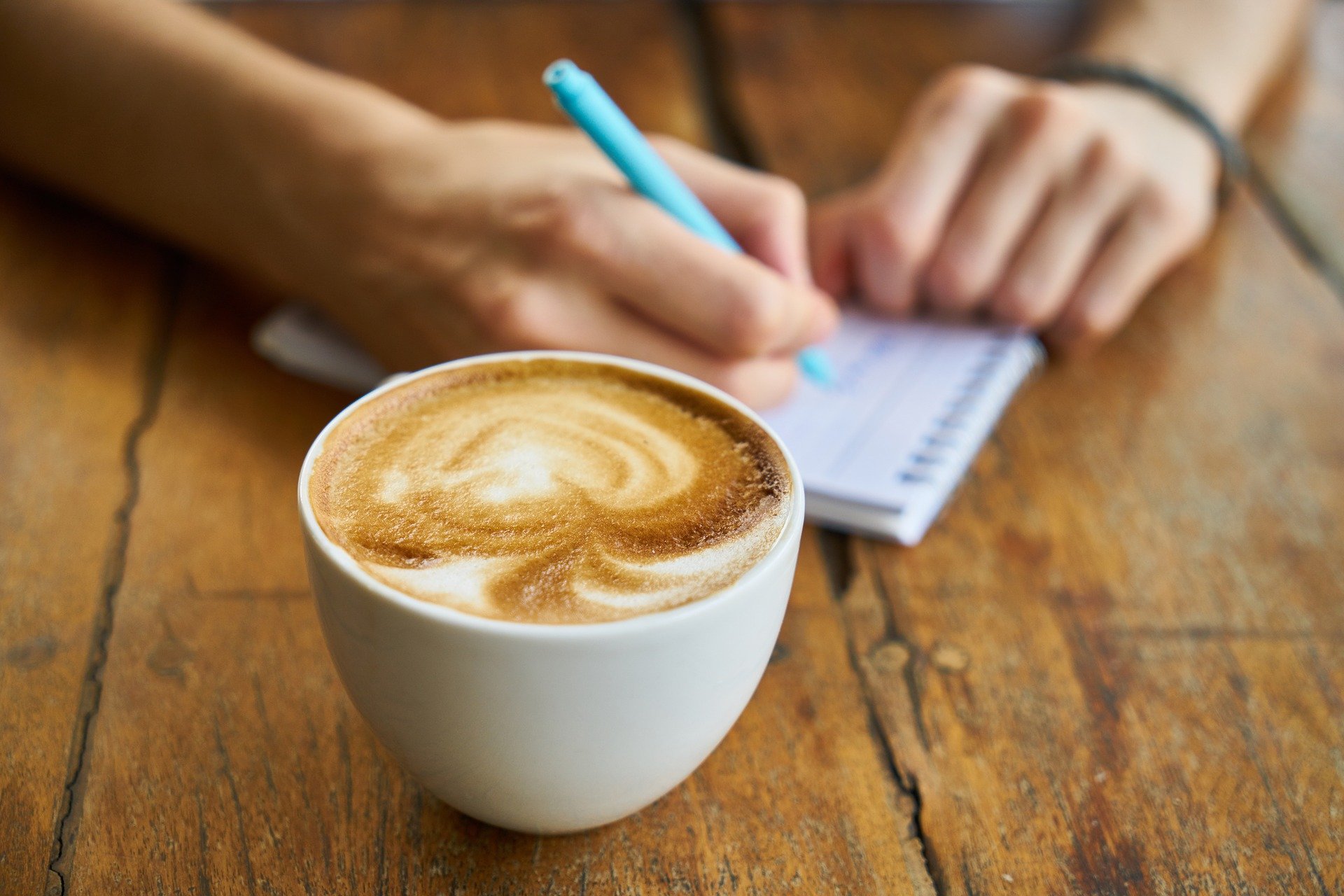 Close up of a cup of frothy coffee on a wooden table. Behind the cup, a pair of hands writes notes on a notepad with a blue pen.