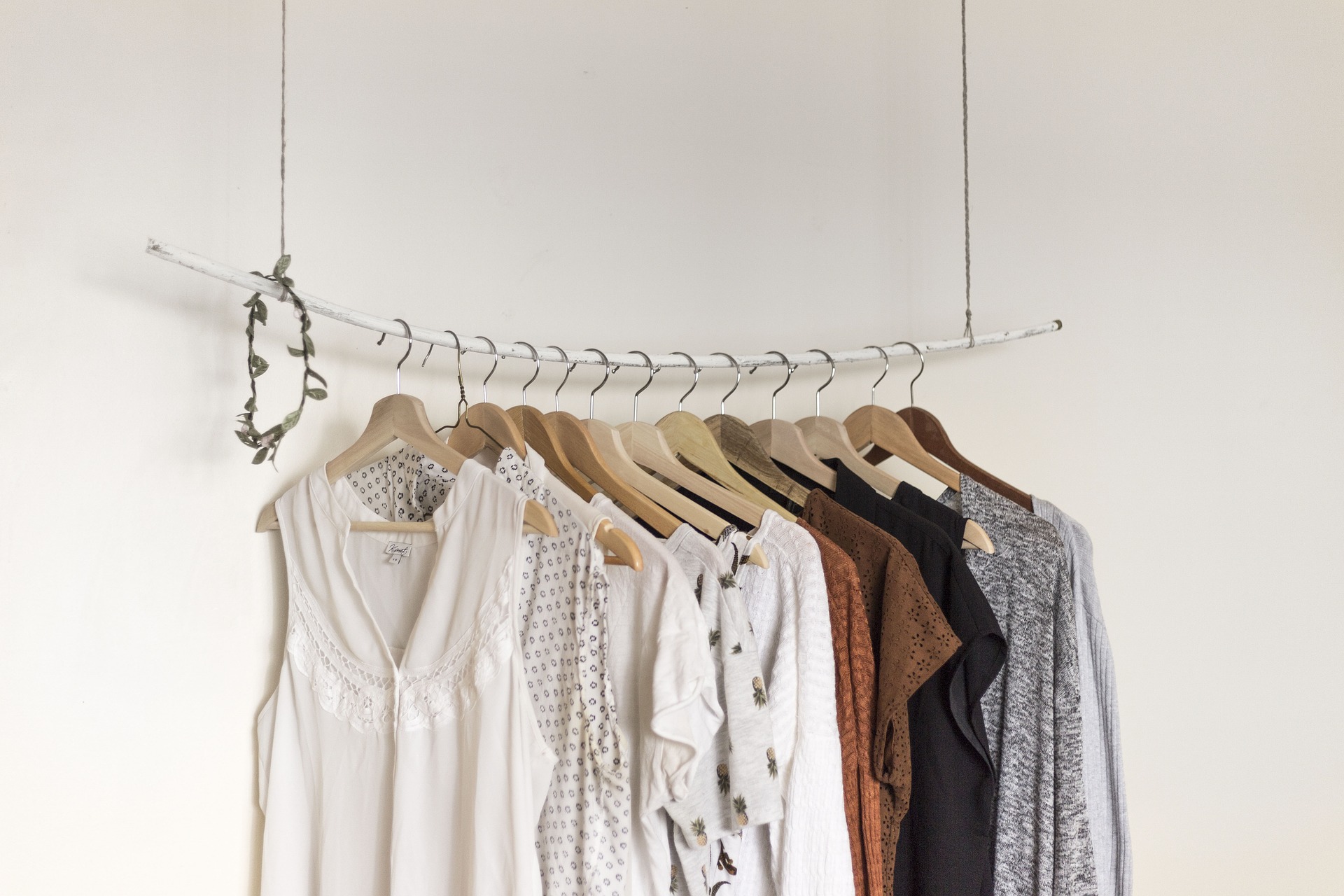 A white bar is suspended on wire against an off-white wall. On the bar hand ladies' blouses in shades of white, brown and black. Some have small prints. A small, green wreath hangs on the left end of the bar.
