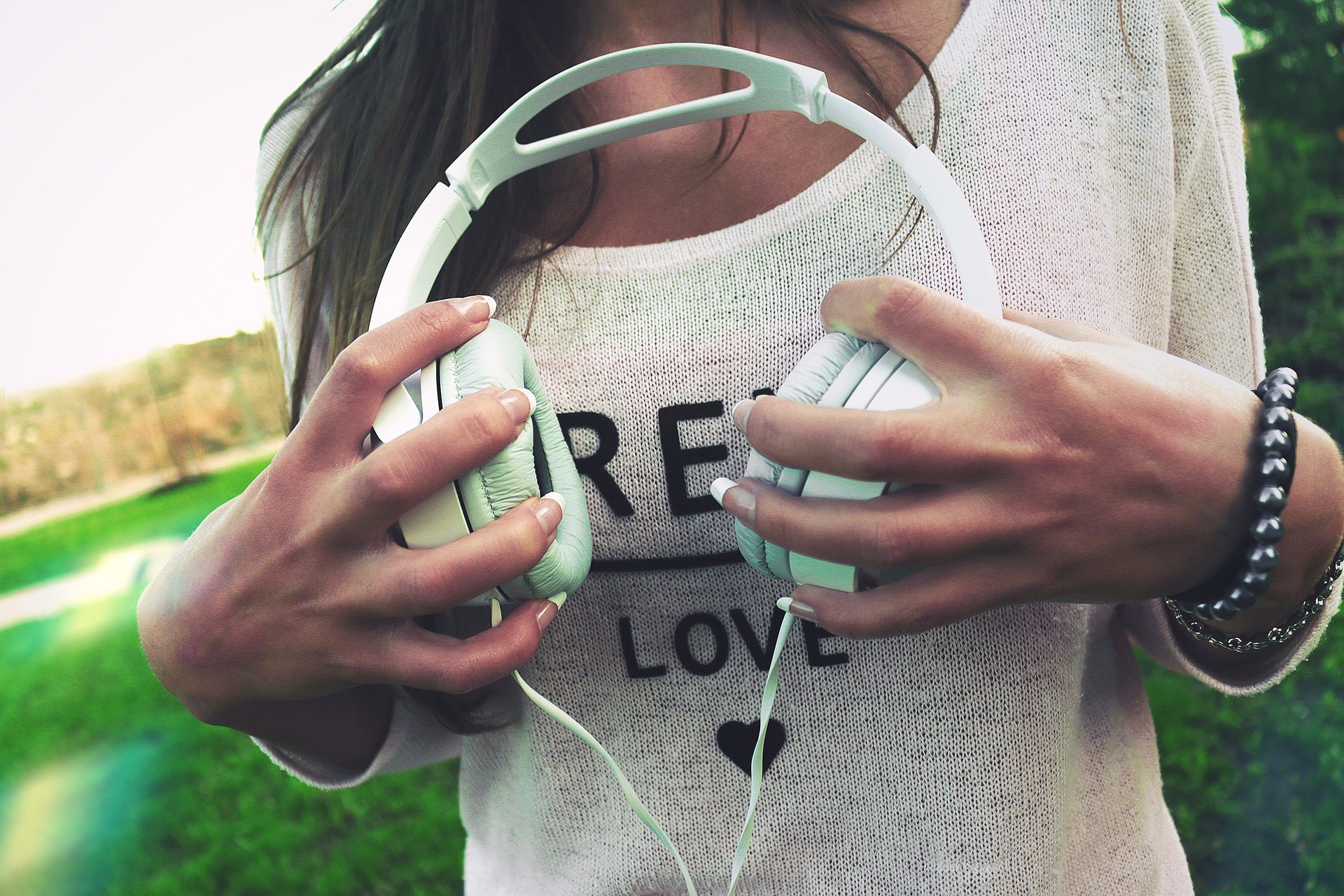A woman stands outside. She holds a pair of mint green headphones in front of her chest. She wears a beige jumper with the word "love" and a black pearl bracelet. 