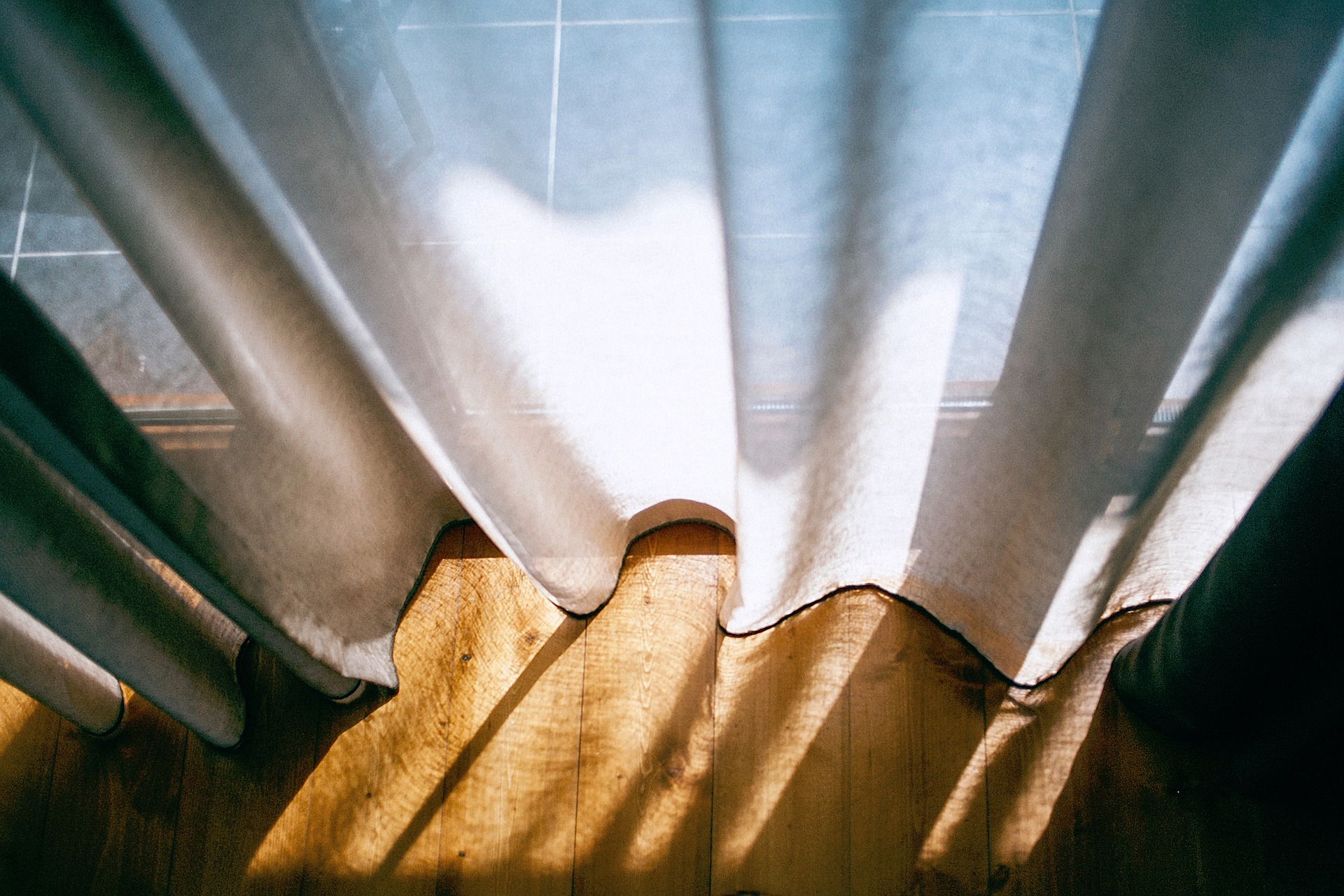 A light, white cotton curtain through which sunshine and light shine, reflecting on a wooden floor.