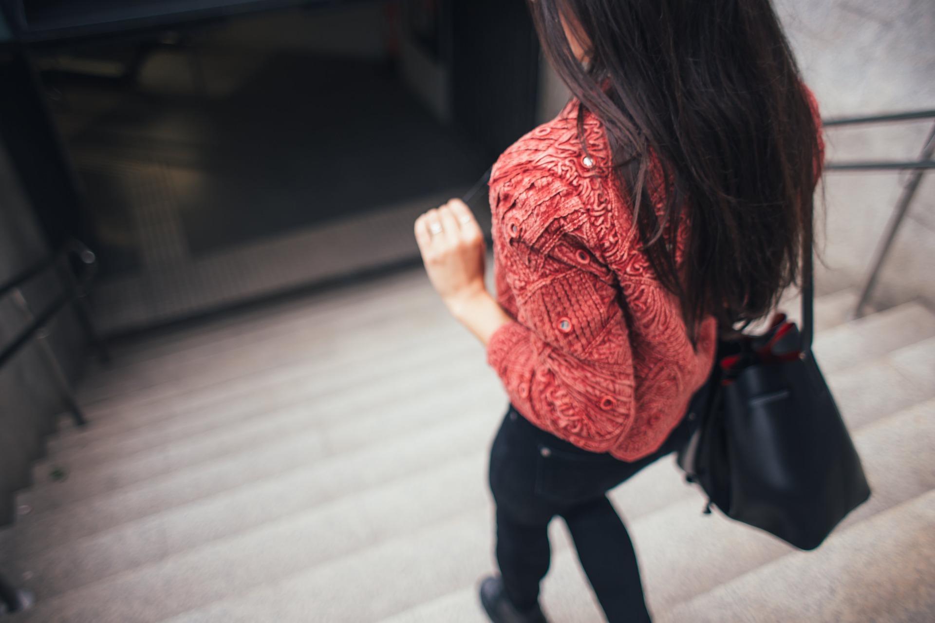 From behind, a woman outside with long, dark hair wearing a rusty-coloured jumper and black trousers walks down concrete stairs. She carries a black handbag over her right shoulder.