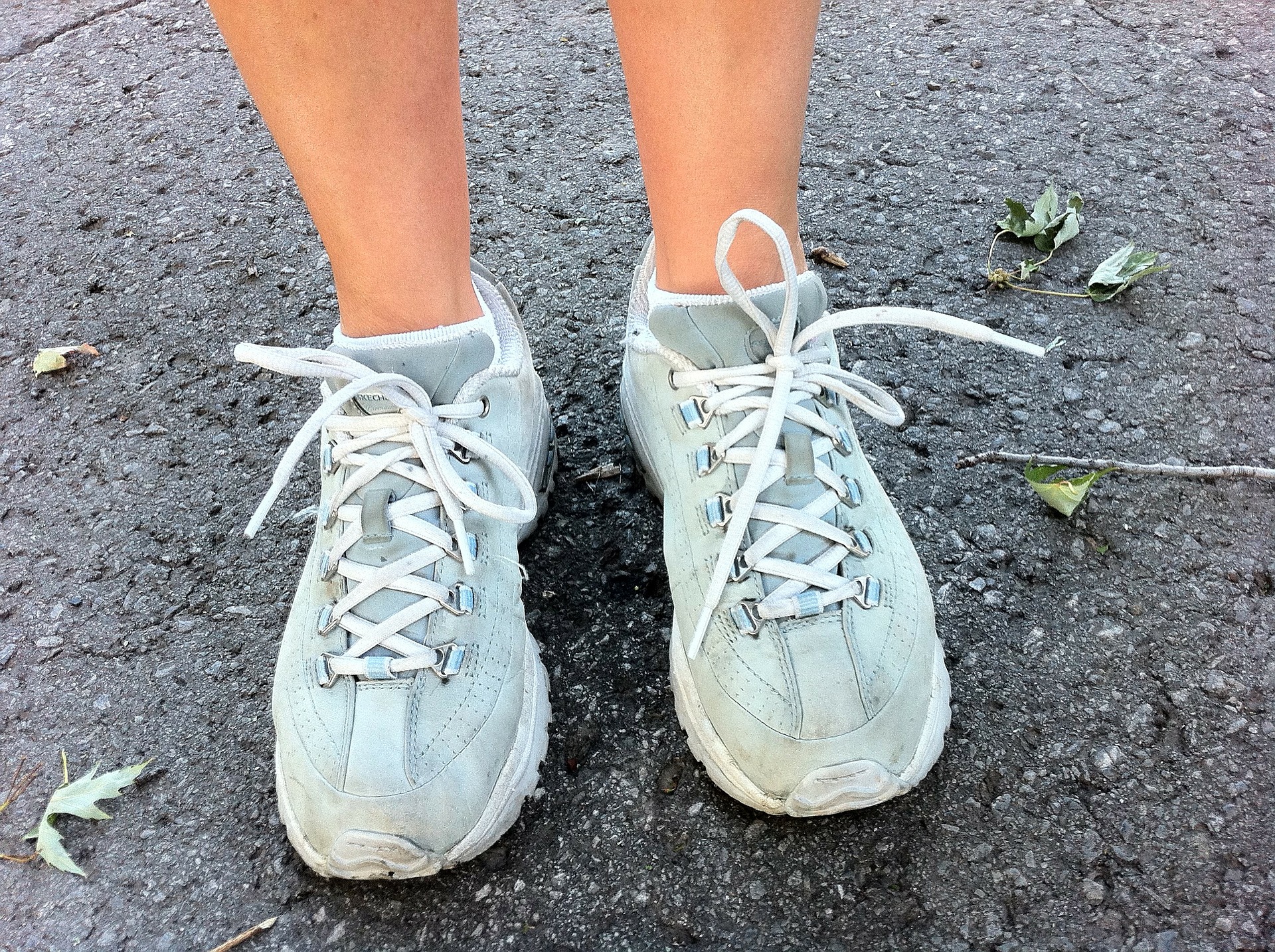 close up of a pair of feet in light blue running shoes and bare ankles. They stand outside on concrete and there are a few fallen leaves.