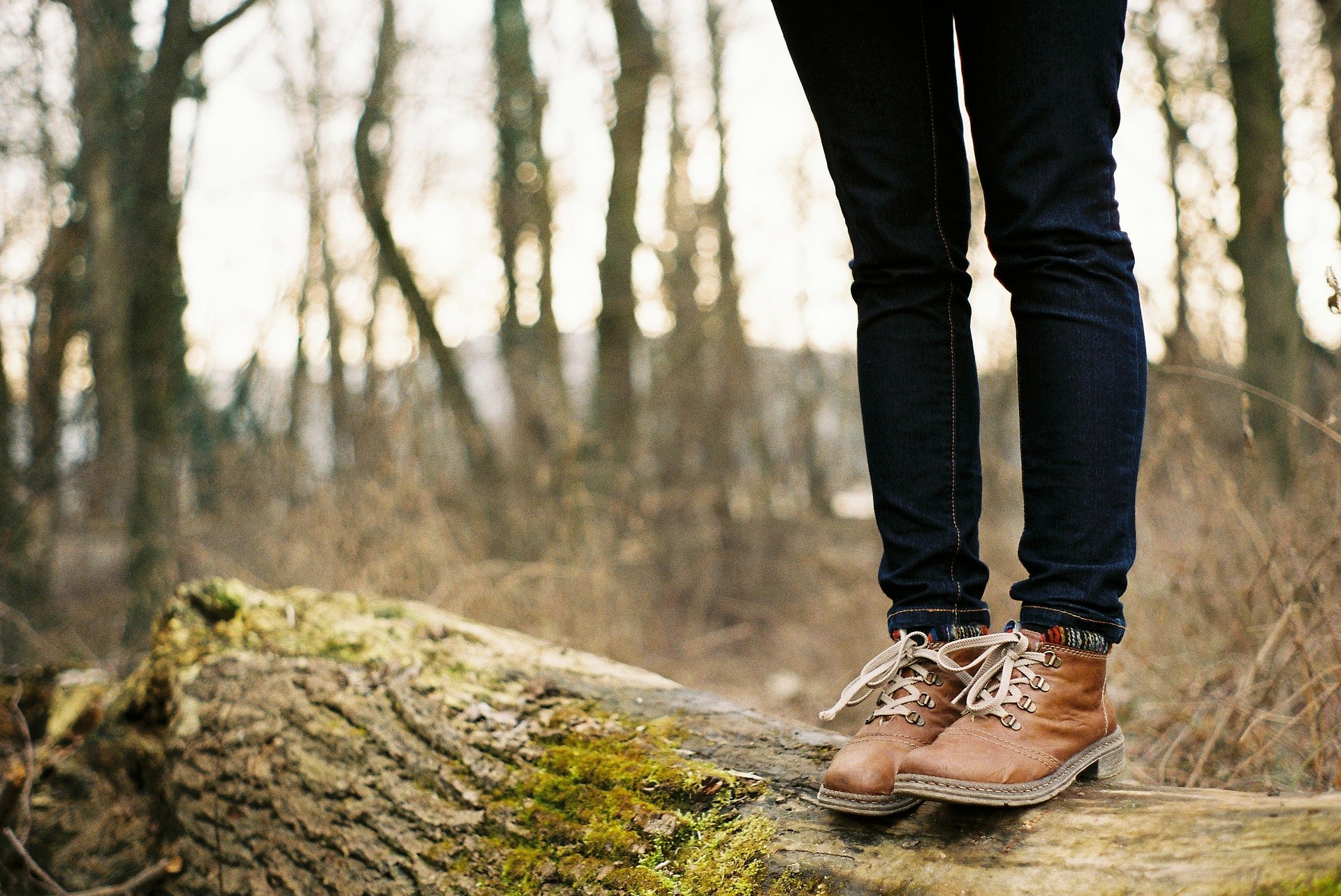 A person - shown from the waist down - stands on a fallen tree trunk covered in moss. They wear balck jeans and brown boots. In the background is a forest and undergrowth.
