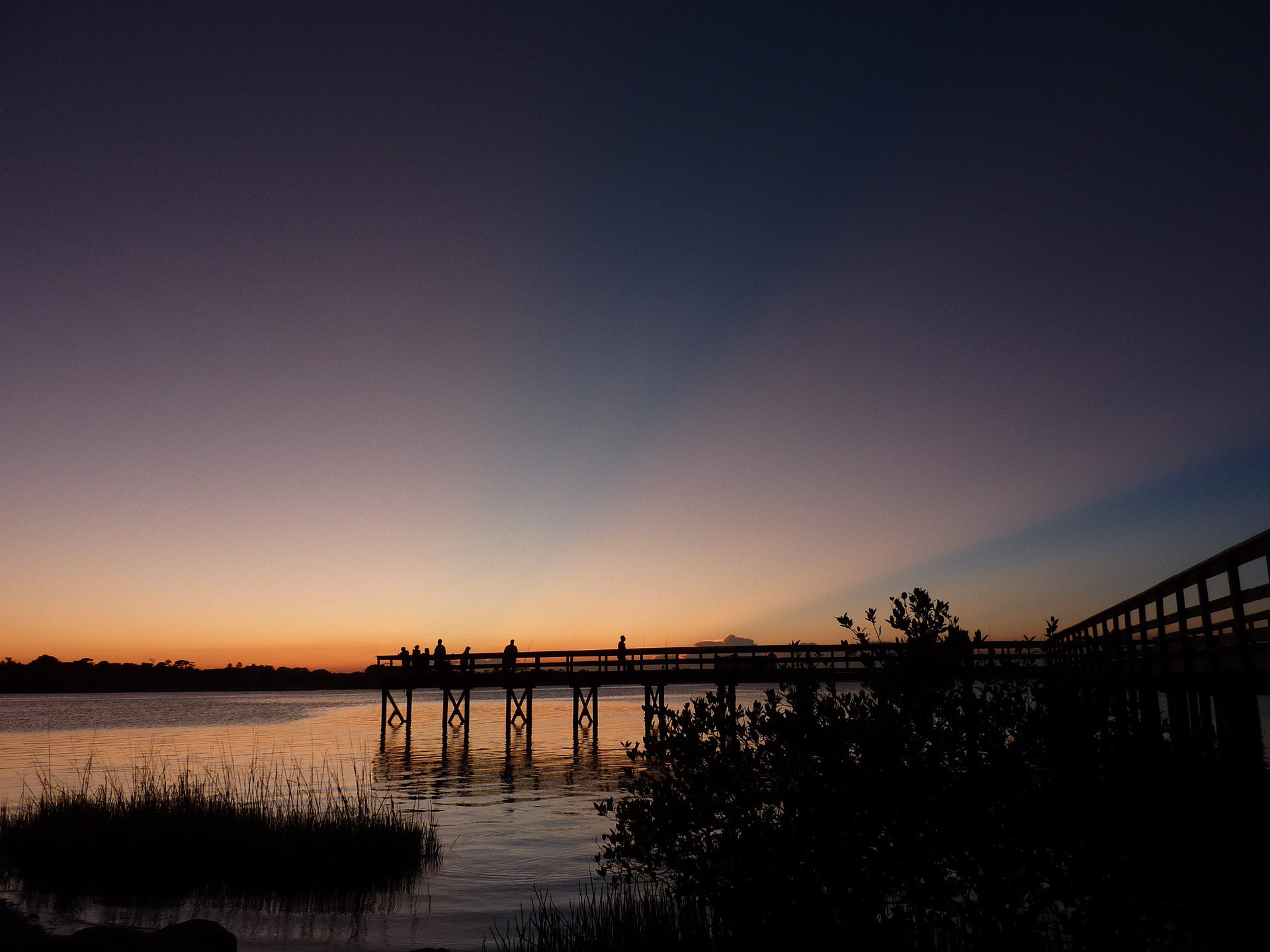 A dock extends into the water at sunset. There are people silhouetted on the dock and water below them reflects a peach and purple coloured sunset