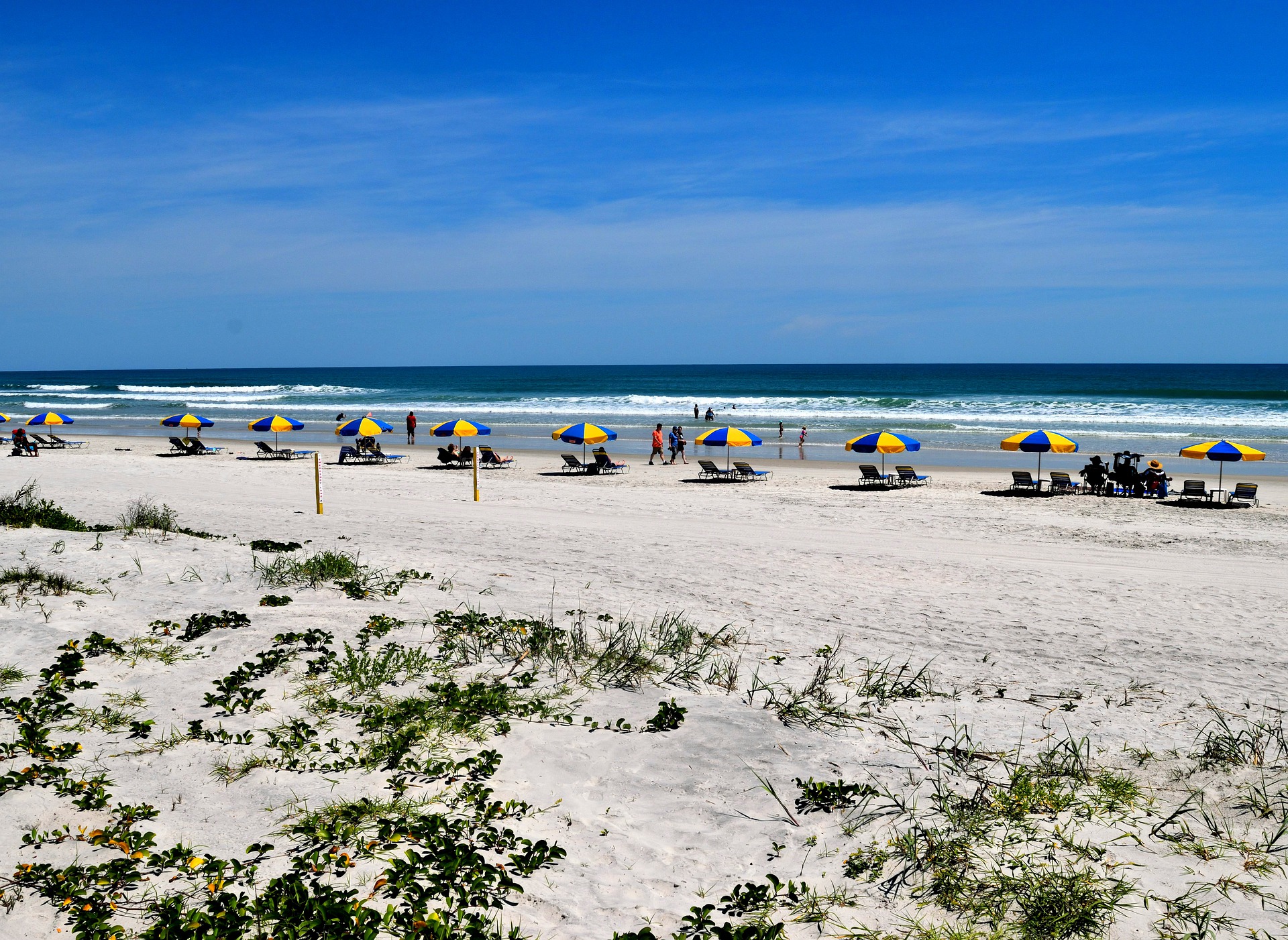 Daytona Beach in Florida. White sand with grasses extends to a blue sea, There are people seated under blue and yellow parasols on the beach and the sky is blue