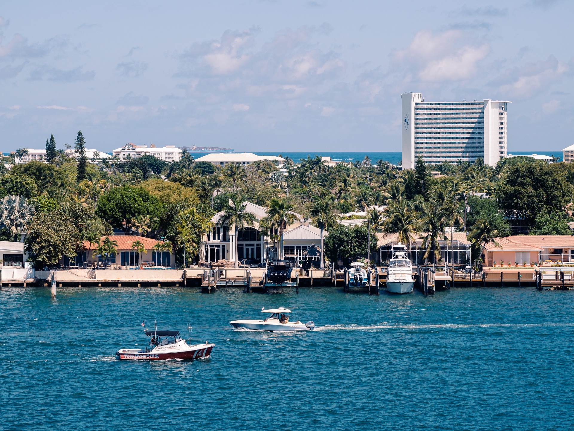 Small yachts in blue water in front of a large white building surrounded by palm trees. On the horizon, there is the sea and other white buildings.