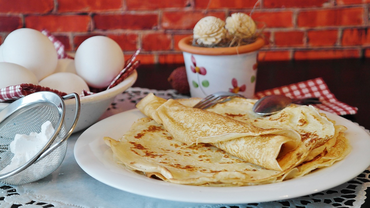 A stack of crepes lay on a white plate. Beside them is a bowl of white eggs. To the left foreground is a small icing sugar sieve.