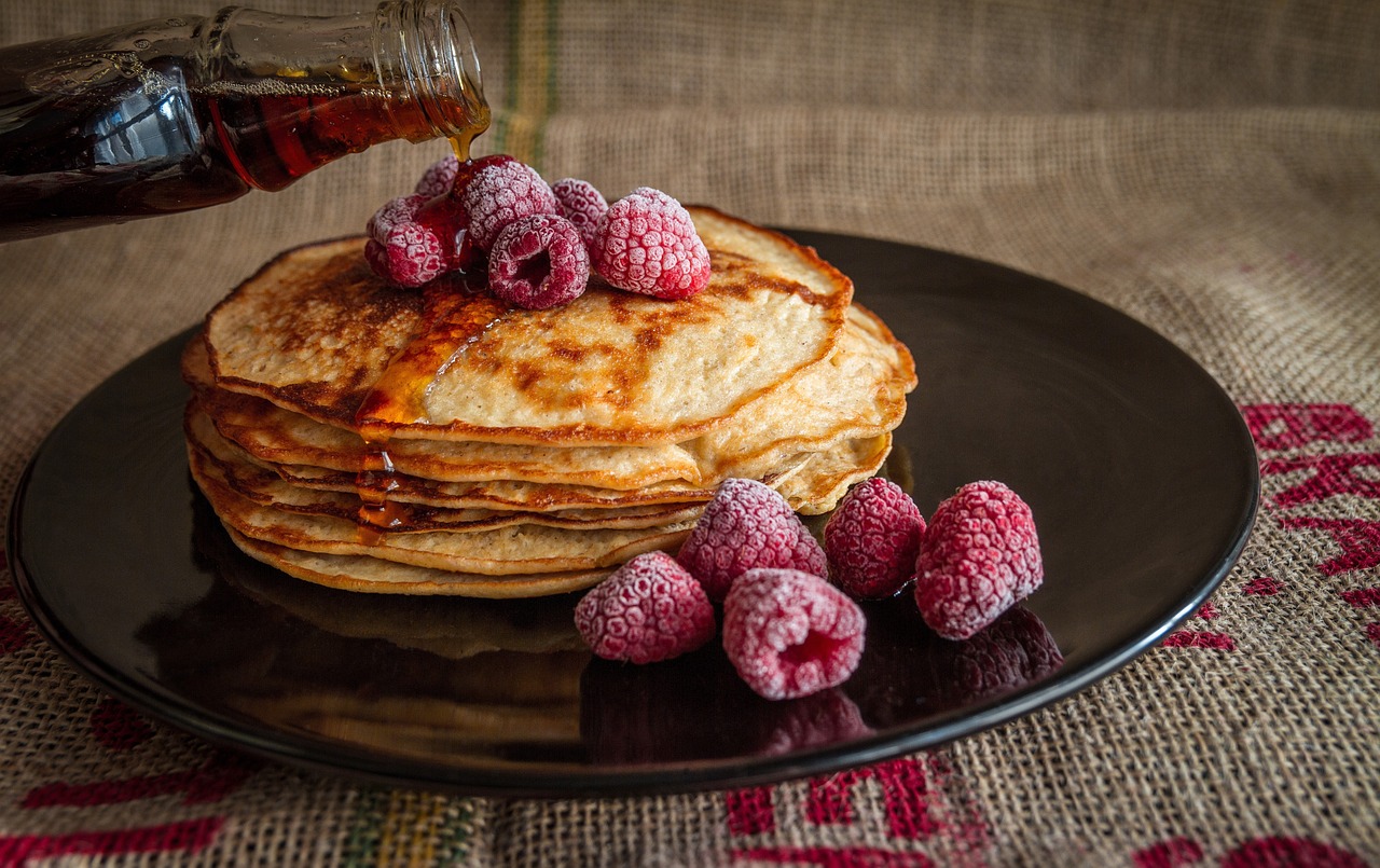 A stack of pancakes are arranged on a large black plate. They are topped with sugar-dusted raspberries. There are more raspberries to the side of the pancakes. Maple syrup is being poured over the top.